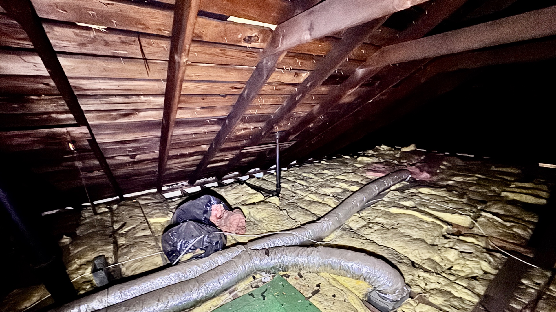 Attic view of skip sheathing with wide intentional gaps between deck boards, alongside old fiberglass insulation and metal ductwork on the attic floor.