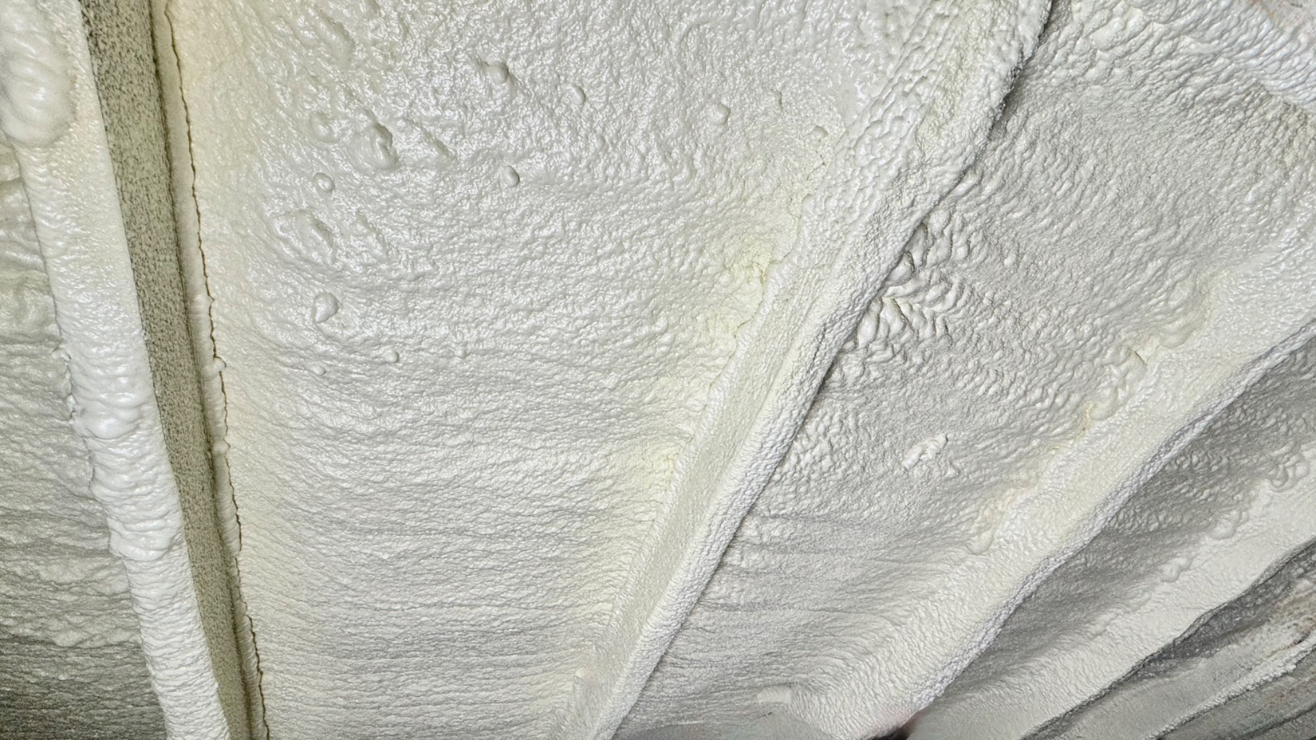Close-up texture of closed-cell polyurethane foam adhesive with a flash coat sealing the sides of a rafter, showing the continuous foam layer.
