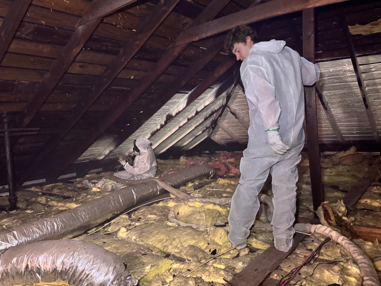 Diversified Energy technician in a full white protective suit standing in an attic while a second technician sprays closed-cell polyurethane foam adhesive onto the underside of the roof deck.
