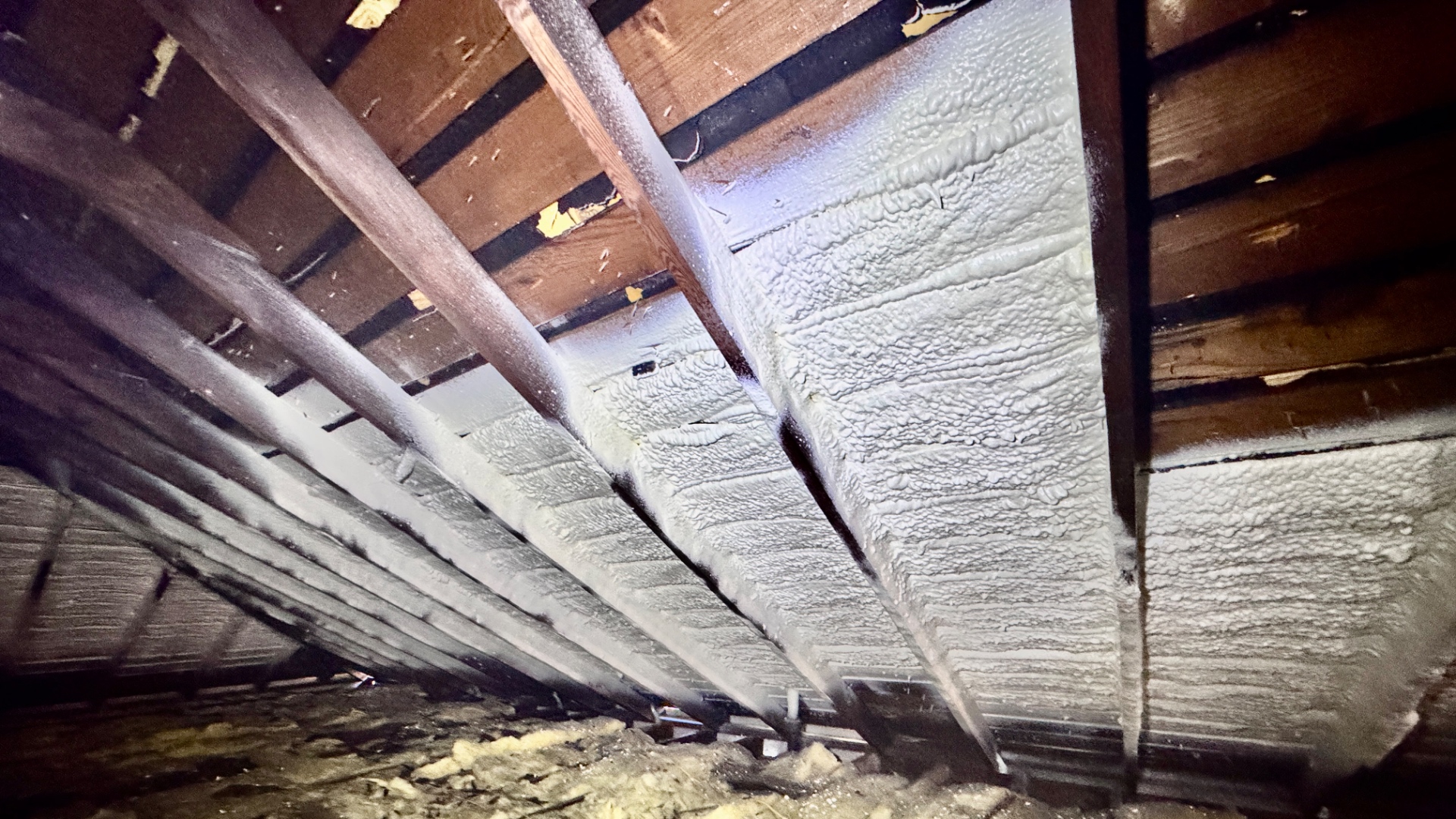Attic view showing bare skip-sheathing on one side and the same deck coated in closed-cell spray foam on the other, illustrating the direct contrast.