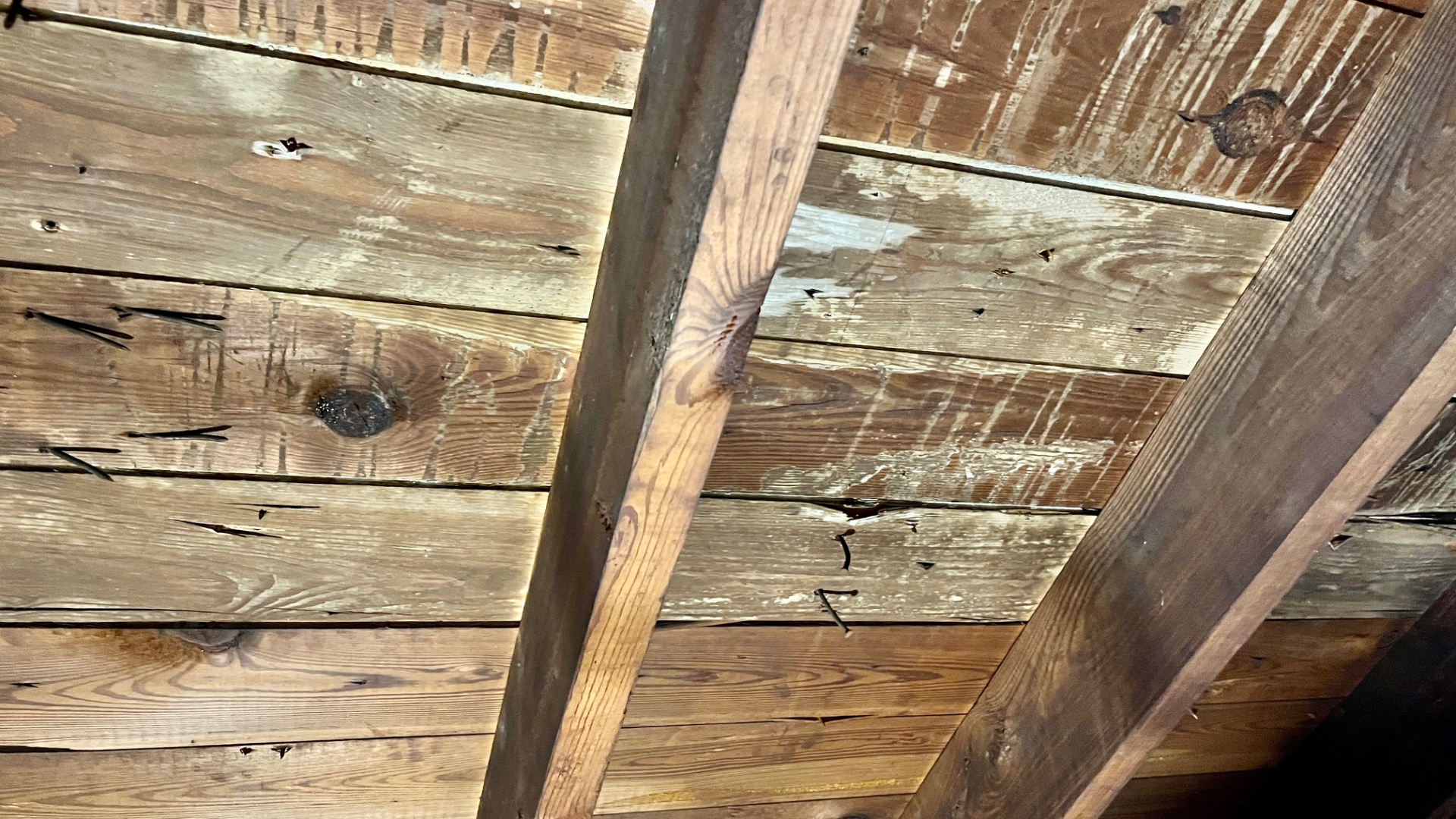 Underside of an old 1x6 plank roof deck showing wood grain, knots, and visible gaps between boards. Nails from shingles above are visible through the boards.