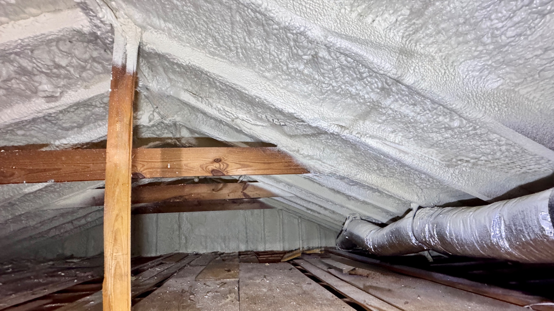 Attic view of a plank-decked roof after closed-cell polyurethane foam adhesive has been sprayed across the entire underside of the deck, with HVAC ductwork now inside a semi-conditioned attic space.